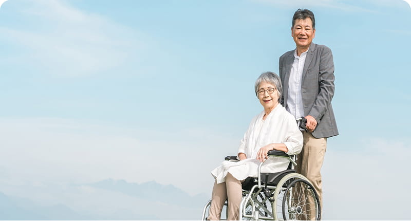 車椅子に座った高齢の女性と、その後ろで笑顔を見せる男性が並んでいる